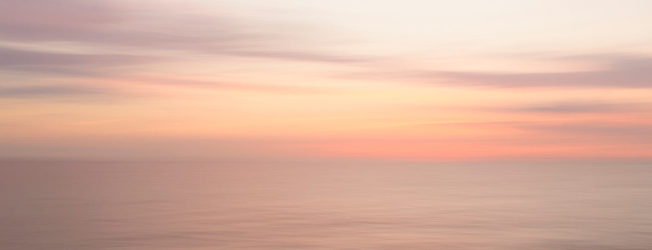 Blurry pastel sunset over calm ocean water with soft pink, orange, and purple tones in the sky.