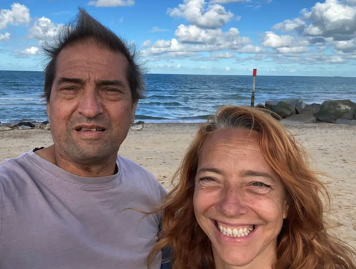 Smiling couple taking a selfie on a sandy beach with the sea and clouds in the background.