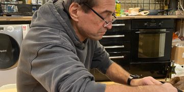 A man in a gray hoodie works on a small electronic device at a cluttered kitchen table filled with tools, dishes, and household items.
