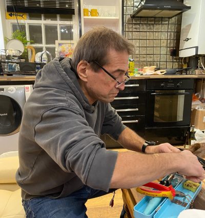 A man in a gray hoodie works on a small electronic device at a cluttered kitchen table filled with tools, dishes, and household items.